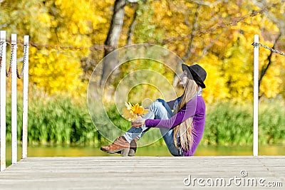 Thoughtful girl sitting on the dock with autumn leaves in hands. Sunny day Stock Photo