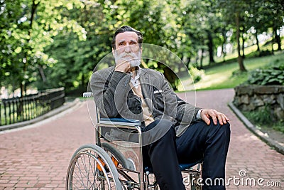 Thoughtful handsome senior bearded disabled man sitting in wheelchair walking in a city park. Concept of disabled people Stock Photo