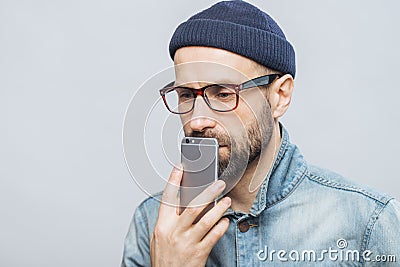 Thoughtful middle aged male with stubble holds smart phone near mouth, being deep in thoughts, thinks about future plans, isolated Stock Photo