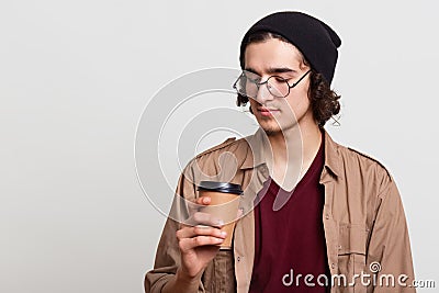 Thoughtful pensive yougster having papercup of coffee, holding hot drink in one hand, looking attentively at it, posing Stock Photo