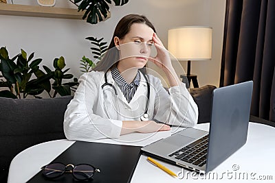 Thoughtful sad young woman doctor sit at work desk with pc laptop, pondering problem solution, pensive therapist physician wearing Stock Photo
