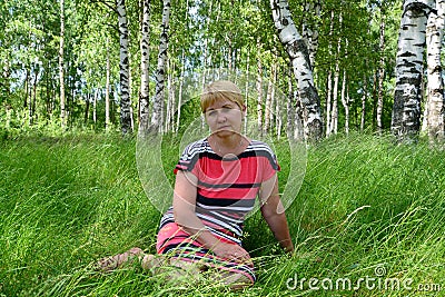 The thoughtful woman of average years sits on a grass in the birch wood Stock Photo