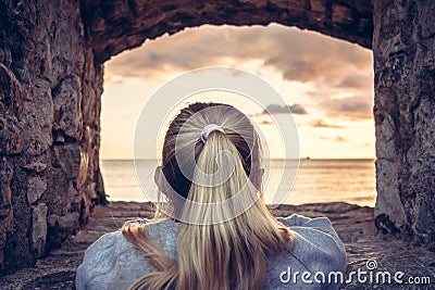 Thoughtful woman devoted into contemplation of beautiful sunset over sea through window of old castle with dramatic sky and perspe Stock Photo