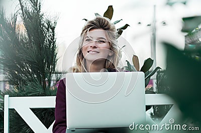 Thoughtful young woman in glasses using a computer, sitting on a bench in a city park. The concept of time is online and work in Stock Photo