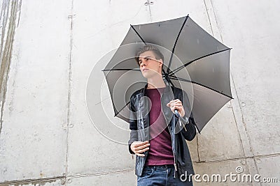 Thougtful young teen man standing in rain in city with umbrella Stock Photo