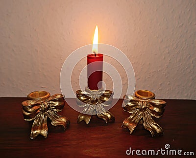 .Three antique golden candlesticks decorated with bows on a mahogany shelf in front of a bright wall. In one is a burning red cand Stock Photo