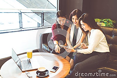 Three beautiful Asian girls using smartphone and laptop, chatting on sofa at cafe Stock Photo
