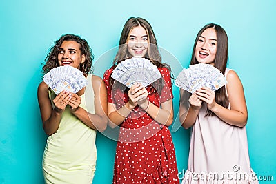 Three beautiful excited three mixed race women in dresses holding money banknotes isolated over blue background Stock Photo