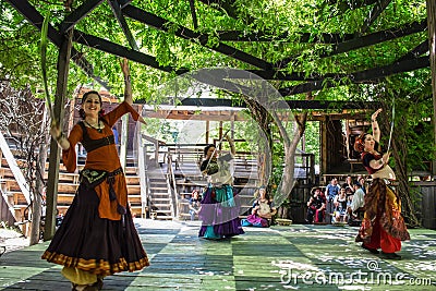 Three belly dancers with swords perform on wooden platform under vined trellis for onlookers in Editorial Stock Photo