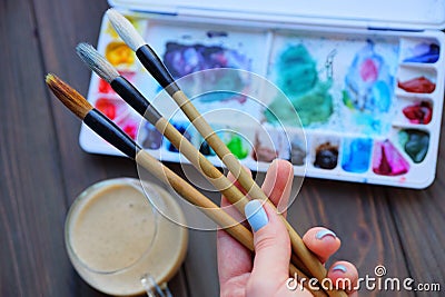 Three brushes in the artist`s hand on the background of a box with colored paints and a glass cup Stock Photo