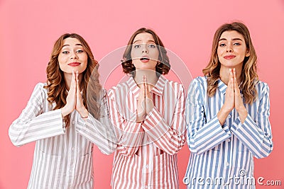 Three concentrated young girls 20s wearing colorful striped pajamas praying keeping palms together and wishing good luck during s Stock Photo