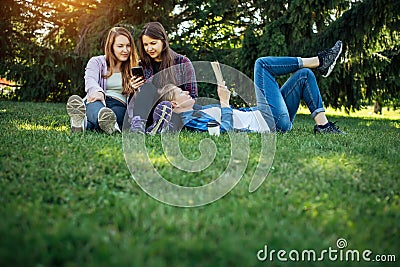 Three cute girls relax and socialize on the lawn in the summer park. Young women on the green grass among the trees, looking Stock Photo