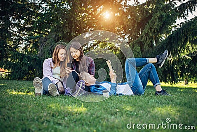 Three cute girls relax and socialize on the lawn in the summer park. Young women on the green grass among the trees, looking Stock Photo