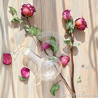 Three dried pink roses, scattered flower petals, green leaves, glass vase on wooden background top view close up Stock Photo