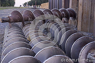 Three earth drills lie side by side on gravel in front of a sheet pile wall at a construction site, one earth drill lies across Stock Photo