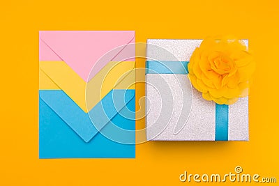 Three envelopes of yellow, blue and pink colors nested in each other lie on a yellow background next to a shiny gift box with a ye Stock Photo