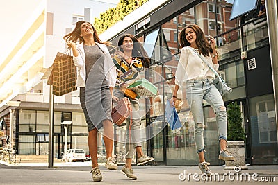 Three fashionable young women strolling with shopping bags. Satisfied women jumping on street. Stock Photo