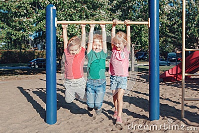 Three funny Caucasian friends hanging on pull-up bars in park on playground. Summer outdoors activity for kids. Active children Stock Photo