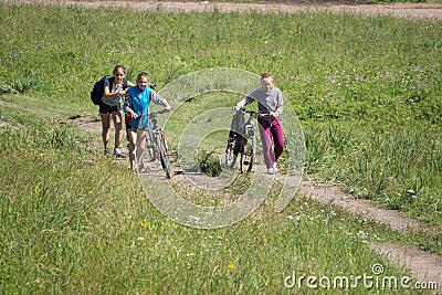 Three girls climb the rural road up the hill with bicycles, among the green grass Editorial Stock Photo