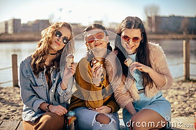 Three girls sit on the beach , near the river, talk, gossip, joke, laugh, smile at womans day. Girls on a hot day eat ice cream. Stock Photo