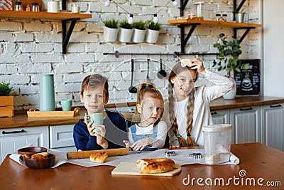 Three happy little kids, siblings cooking together, rolling out dough, standing at the wooden countertop in the modern kitchen, Stock Photo