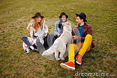 A three happy young stylish friends spend time outdoors together with their husky dog sitting on green grass. Stock Photo