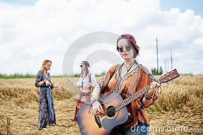 Three hippie women, wearing boho style clothes, dancing in the wheat field, playing guitar, laughing, Female friends, traveling Stock Photo