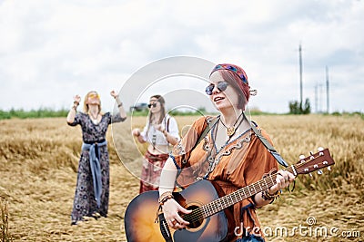 Three hippie women, wearing boho style clothes, dancing in the wheat field, playing guitar, laughing, Female friends, traveling Stock Photo