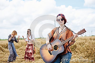 Three hippie women, wearing boho style clothes, dancing in the wheat field, playing guitar, laughing, Female friends, traveling Stock Photo