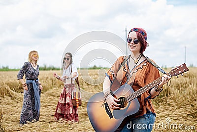 Three hippie women, wearing boho style clothes, dancing in the wheat field, playing guitar, laughing, Female friends, traveling Stock Photo