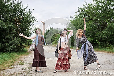 Three hippie women, wearing boho style clothes,, walking on dirt road in countryside, dancing, relaxing, having fun. Friends, Stock Photo