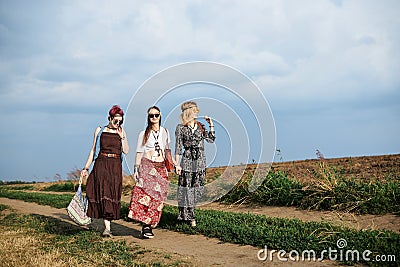 Three hippie women, wearing boho style clothes, walking on dirt road on green field, having fun. Female friends, traveling Stock Photo