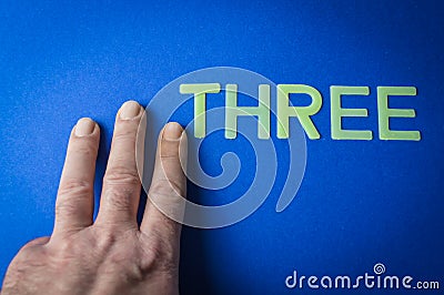 Three human fingers beside the word Three written with plastic letters on blue paper background Stock Photo
