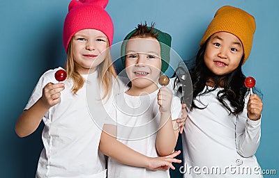 Three joyful friends kids in white t-shirts and colorful hats hold sweet lollipop candies happy smiling hugging Stock Photo