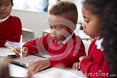 Three kindergarten school kids sitting at desk in a classroom using a tablet computer and stylus together, selective focus Stock Photo