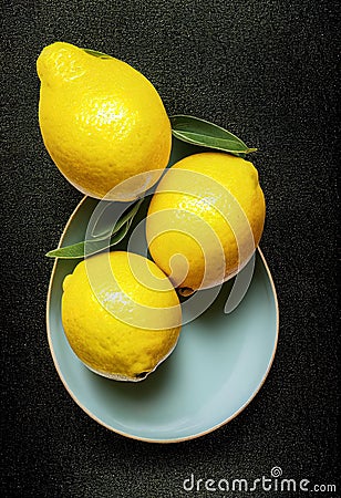 three lemons on a plate with a green leaf on top of it on a black surface with a black background Stock Photo