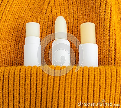 Three lip balms on the orange knitted background pocket. Winter lip care sticks with beeswax, honey, panthenol and shea butter. Stock Photo