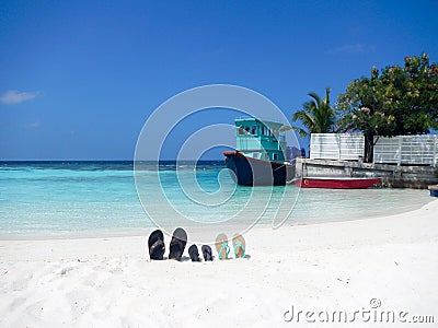 Three pairs of flip flops sandals on a Maldive beach propped up in the sand with an old fishing boat at a quay behind and palm tre Editorial Stock Photo