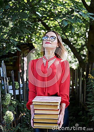 Three-quarter portrait of young blond woman, wearing eyeglasses, holding a bunch of books. Student with books in the park. Stock Photo