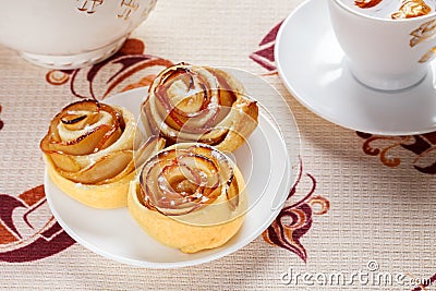 Three ruddy buns on a white saucer with herbal apple tea in a cup and a teapot in the background. Stock Photo