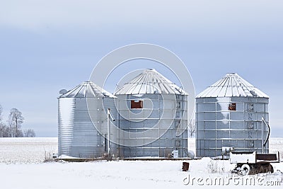 Three steel silos or grain bins covered in snow on a farm in a rural area in winter with a small wagon in the foreground Stock Photo