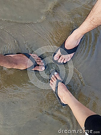 Three tourists getting their feet wet with slippers together in sea water on sandy beach Stock Photo