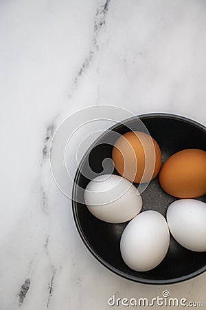 Three white Organic Eggs with Two Organic Brown Egg in a black Bowl on a Marble Counter Top Stock Photo