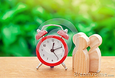 Three wooden human figures standing next to a red alarm clock on a nature background. Time concept. Business, Hourly payment Stock Photo