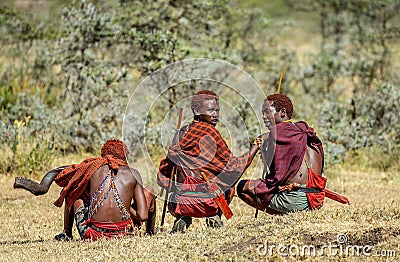 Three young Masai warriors in traditional clothes and weapons are sitting in the savannah. Editorial Stock Photo