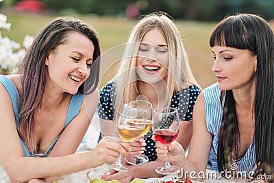 Three young women in blue dresses, and hats lie on plaid and drink wine. Outdoor picnic on grass on beach. Delicious Stock Photo