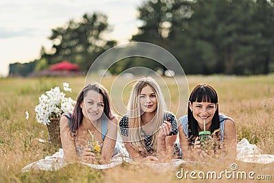 Three young women in blue dresses, and hats lie on plaid and drink wine. Outdoor picnic on grass on beach. Delicious Stock Photo