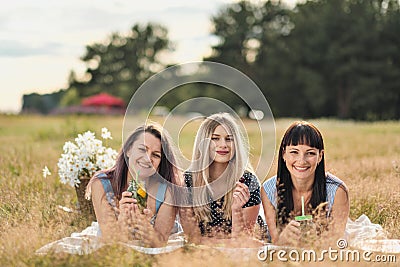 Three young women in blue dresses, and hats lie on plaid and drink wine. Outdoor picnic on grass on beach. Delicious Stock Photo