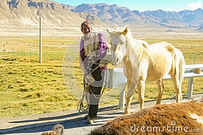 CLOSE UP: Old Tibetan farmer leads beautiful albino horse down an asphalt road. Editorial Stock Photo