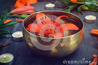 Tibetan singing bowl with floating lily inside. Burning candles, lily flowers and petals on the black wooden background. Meditatio Stock Photo